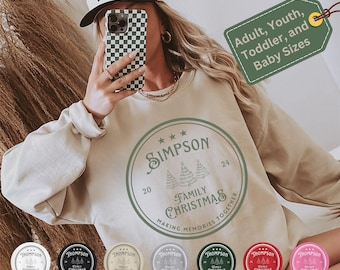 Könnte beinhalten: Ein beiger Pullover mit einem grünen Kreisdesign, auf dem "SIMPSON FAMILY CHRISTMAS MAKING MEMORIES TOGETHER" steht, mit drei stilisierten Weihnachtsbäumen im Kreis. Der Pullover ist in den Größen Erwachsener, Jugendlicher, Kleinkind und Baby erhältlich.