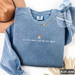 May include: Blue jean Comfort Colors sweatshirt featuring the text "AIN'T NO GHOST LIKE THE HOLY GHOST" and a small ghost graphic. A gold necklace and a brown hat are visible in the background.
