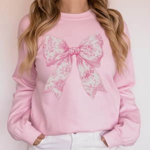 May include: A pink sweatshirt with a large white floral bow design printed on the front.