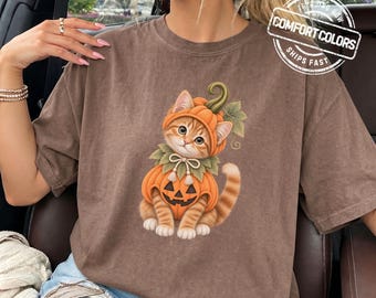 May include: A brown short-sleeved t-shirt featuring a graphic of an orange tabby kitten wearing a pumpkin costume with a green stem hat and a jack-o'-lantern body. The shirt includes a circular logo in the top right corner with the text COMFORT COLORS.