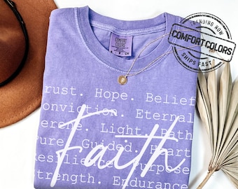 May include: A lilac Comfort Colors t-shirt with the word "Faith" in a large, stylized font. The shirt features additional words like "Hope", "Belief", and "Endurance". A gold necklace and a brown hat are also visible.
