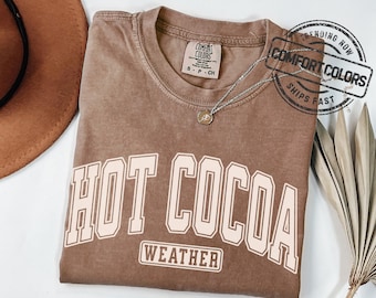 May include: Espresso-colored t-shirt with the words "HOT COCOA WEATHER" in a cream-colored, collegiate-style font. The shirt has a crew neck and a small tag at the neckline. A silver necklace with a pendant is draped on the shirt.
