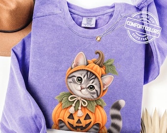 May include: A violet long-sleeve sweatshirt featuring a graphic of a gray tabby kitten wearing a pumpkin costume with a jack-o'-lantern face. The garment has a Comfort Colors brand label at the inner neckline and a crew neck collar design.