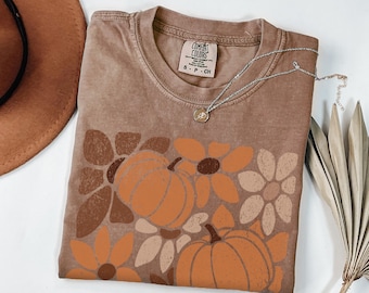 May include: Brown t-shirt with a pumpkin and floral design. The shirt has a crew neck and short sleeves. A silver necklace with a small pendant rests on the shirt. The shirt is labeled "Comfort Colors".