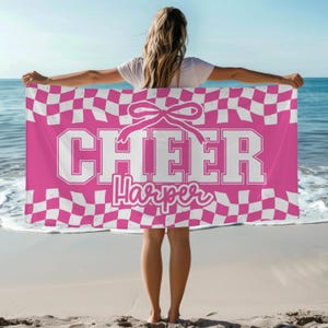 May include: A pink and white checkered beach towel with the word "CHEER" in large white block letters and the name "Harper" in a script font. A white bow is above the word "CHEER". The towel is held up on a beach with the ocean in the background.