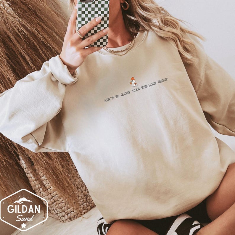 May include: A beige sweatshirt featuring the phrase "Ain't No Ghost Like The Holy Ghost" in black text, with a small ghost graphic. The sweatshirt is a Gildan Sand brand. The sleeves are long and the fit is relaxed.