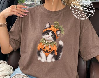 May include: A brown short-sleeve t-shirt featuring a graphic of a black and white kitten wearing a pumpkin costume with a green stem and leaves. The kitten is centered on the chest. A circular logo in the top right corner reads COMFORT COLORS.