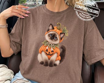 May include: A brown short-sleeve t-shirt featuring a graphic of a Siamese cat wearing a pumpkin costume with a jack-o'-lantern face and green leaves. The shirt has a crew neckline and a relaxed fit. The text COMFORT COLORS is visible.