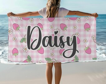 May include: A pink and white gingham beach towel with strawberries, daisies, and the name "Daisy" in black script. The towel is held up on a beach with the ocean in the background.