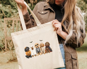 May include: A cream-colored canvas tote bag featuring a graphic print of five dogs in Halloween costumes, including a pumpkin, a ghost, a skeleton, a mummy, and a vampire, accompanied by two small black bat illustrations above the dogs.