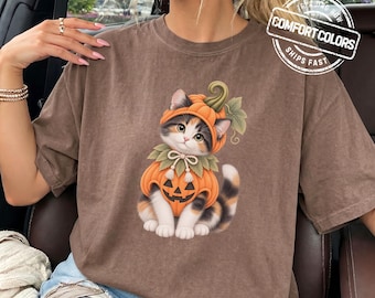 May include: A brown short-sleeved t-shirt featuring a graphic of a calico kitten wearing a pumpkin costume with a stem hat and a jack-o'-lantern body. The shirt has a round neckline and a relaxed fit. The text ESPRESSO is printed in the corner.