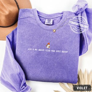 May include: A violet Comfort Colors sweatshirt with the embroidered text "AIN'T NO GHOST LIKE THE HOLY GHOST" below a small ghost graphic. The sweatshirt has a crew neck and long sleeves. A brown hat and a gold necklace are visible.