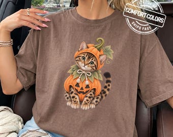 May include: A brown short-sleeved t-shirt featuring a graphic of a tabby kitten wearing a pumpkin costume with a green stem and leaves. The kitten has a bow tie and a carved jack-o'-lantern face on the pumpkin body. The shirt has a crew neckline.