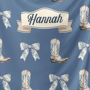 May include: A blue fabric with a white ribbon that says "Hannah" in a black script font. The fabric is decorated with a repeating pattern of white cowboy boots with blue floral designs and white bows with blue floral designs.