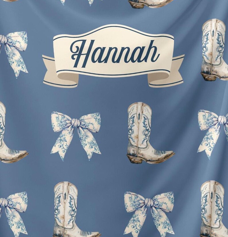 May include: A blue fabric with a white ribbon that says "Hannah" in a black script font. The fabric is decorated with a repeating pattern of white cowboy boots with blue floral designs and white bows with blue floral designs.