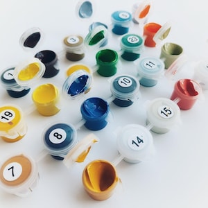 Op de afbeelding: Een verzameling kleine, ronde plastic verfpotjes met bijbehorende deksels, elk voorzien van een uniek nummer. De potjes bevatten verschillende kleuren acrylverf, waaronder tinten geel, blauw, groen, rood, bruin en wit, gerangschikt in een groep.