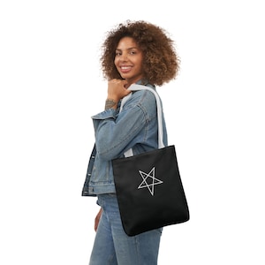 May include: A black tote bag with a white pentagram design. The bag has white straps and is being held by a person wearing a denim jacket and jeans.