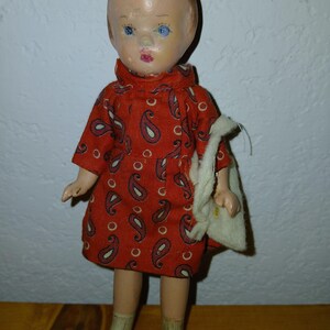 May include: A vintage doll with a painted face, blue eyes, and light brown hair, wearing a short-sleeved red dress featuring a dark blue and white paisley pattern. The doll has light-colored skin, wears white socks, and black shoes, and holds a small white fabric bag.