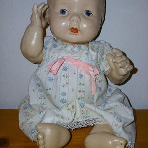 May include: A vintage doll with a molded head, blue eyes, and painted orange lips. The doll wears a white dress featuring a vertical pattern of small blue and pink flowers, accented with a light pink ribbon bow at the chest and white lace trim.