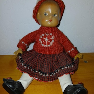 May include: A doll with a tan face and brown hair wearing a red knitted sweater with a white snowflake design and pearl accents, a red and black houndstooth skirt with black bead trim, white leggings, and black shoes with small red buttons.