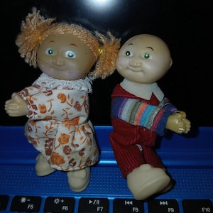 May include: Two small dolls with molded plastic heads and bodies. The left doll has orange yarn hair in pigtails and wears a white dress with an orange floral pattern. The right doll is bald and wears a red outfit with a multi-colored striped long-sleeve shirt.