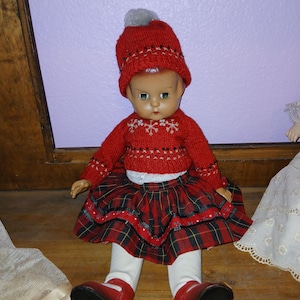 May include: A doll wearing a red knit hat with a gray pom-pom, a red long-sleeved sweater with white snowflake patterns, a red and green plaid skirt with black trim, white leggings, and red shoes with black soles, sitting in a seated position.
