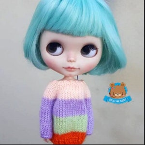 Può includere: Una bambola con un taglio bob turchese e grandi occhi espressivi. La bambola indossa un maglione a righe nei toni del rosa, viola, verde e arancione. È visibile un piccolo logo con un orso e la scritta "DOLLS ARE GOOD".