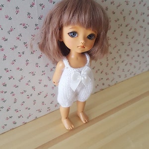 Puede incluir: Muñeca de piel clara, ojos azules y pelo corto castaño claro. La muñeca lleva un pelele blanco de punto con un lazo. El fondo presenta un estampado floral sobre un fondo rosa claro.