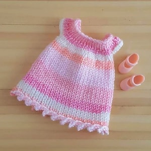 Puede incluir: Un vestido de punto de rayas rosa y blanco con un dobladillo festoneado. El vestido está hecho para una muñeca y tiene un diseño sencillo.