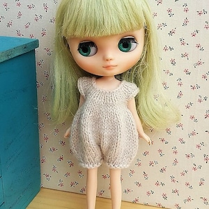 Puede incluir: Una muñeca con cabello verde y ojos verdes grandes que lleva un pelele de punto beige claro. La muñeca está de pie frente a un papel pintado con estampado floral y un pequeño armario azul.