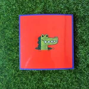 May include: A red square sticker with a blue border featuring a cartoon alligator peeking out of the water. The alligator is green with white teeth and a happy expression.