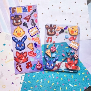 Peut inclure: Une feuille de plastique transparente avec des autocollants colorés représentant des personnages du jeu vidéo "FNAF". Les autocollants comprennent Freddy Fazbear, Chica la Poule, Bonnie le Lapin, Foxy le Renard Pirate et d'autres personnages. Le texte "Let's Eat!!!" apparaît sur deux des autocollants.
