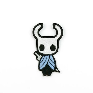 Peut inclure: Un patch brodé noir et blanc représentant le Chevalier, personnage du jeu vidéo Hollow Knight. Le Chevalier porte une cape bleue et noire et tient une épée.