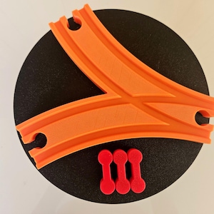 Può includere: Vista dall'alto di una base circolare nera con un pezzo di binario ferroviario in plastica arancione e tre piccoli connettori rossi. Il binario ha un design a forma di Y con sezioni curve e fessure per i connettori.