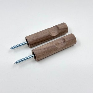 Puede incluir: Dos ganchos de pared de madera con un tornillo en cada extremo. Los ganchos son de madera marrón claro y tienen una forma redondeada.