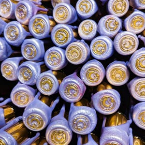 Puede incluir: Primer plano de un grupo de tapones de botella morados y dorados con un diseño de luna creciente. Los tapones están dispuestos en un patrón aleatorio, creando una exhibición visualmente atractiva y única.