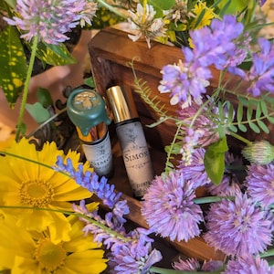 May include: Two perfume bottles with gold caps and labels that read "Simon" are surrounded by purple and yellow flowers. One bottle has a green wax seal with a mushroom design. The bottles are displayed in a wooden box.