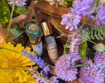 Pode incluir: Duas garrafas de perfume com tampas douradas e rótulos que dizem "Simon" são cercadas por flores roxas e amarelas. Uma garrafa tem um selo de cera verde com um desenho de cogumelo. As garrafas são exibidas em uma caixa de madeira.