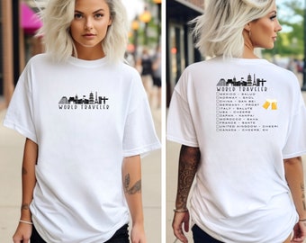 May include: White t-shirt with a black graphic of a city skyline and the text "World Traveler". The back of the shirt has a list of countries and their cheers, such as "Mexico - Salud", "Norway - Skol", and "Germany - Prost".