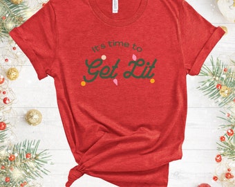 May include: Red t-shirt with the phrase "It's time to Get Lit" in green script. The design includes colorful accents resembling Christmas lights. The shirt is knotted at the bottom and has rolled-up sleeves. The background features Christmas decorations.