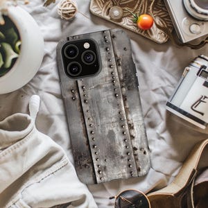 Puede incluir: Funda de teléfono con diseño de metal envejecido. La funda gris tiene un aspecto industrial con remaches visibles y una superficie texturizada. Diseñada para un smartphone moderno con cámara de múltiples lentes.