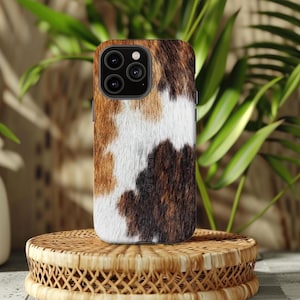May include: A smartphone case featuring a cowhide design in brown, white, and black. The case is placed on a circular, woven stand, with green leaves in the background. The case has a multi-lens camera module.