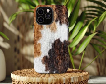 May include: A smartphone case with a cowhide pattern in shades of brown, white, and black. The case is on a woven, circular stand, with green foliage in the background. The case has a camera module with multiple lenses.