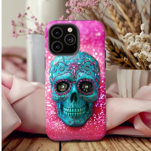 Puede incluir: Una funda de teléfono rosa con purpurina con un diseño de calavera de azúcar azul y blanco. La calavera tiene diamantes de imitación para los ojos y una boca turquesa.