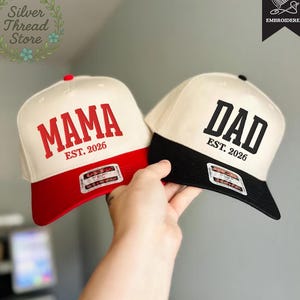May include: Two baseball caps, one red and cream, the other black and cream. The red cap says "MAMA EST. 2026" in red block letters. The black cap says "DAD EST. 2026" in black block letters. Both have a flat brim.