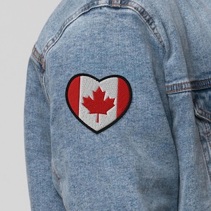 May include: A denim jacket with a heart-shaped patch featuring the Canadian flag. The patch has a red maple leaf on a white background, framed by a red border and a black outline. The jacket is light blue.