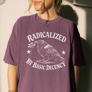 May include: Berry-colored t-shirt with the text "RADICALIZED BY BASIC DECENCY" and a graphic of a raven holding a sprig. The design is in white. The shirt is a short-sleeved, crew neck style.