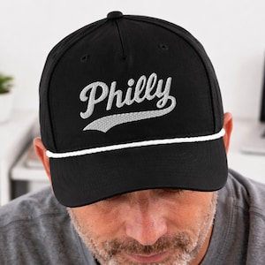 May include: Black baseball cap with the word "Philly" embroidered in white script with a white accent. The cap has a curved brim and a white rope detail around the base. A stylish accessory for any casual outfit.