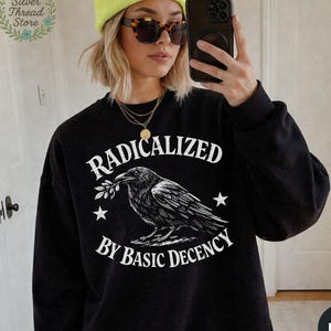 May include: Black sweatshirt with the text "RADICALIZED BY BASIC DECENCY" in white. A detailed illustration of a raven holding a sprig is centered below the word "RADICALIZED". Two stars flank the raven. The sweatshirt is a solid black color.