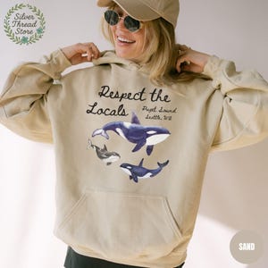 May include: Sand-colored hoodie featuring a graphic of orcas with the text "Respect the Locals" and "Puget Sound, Seattle, WA." The design includes three orcas in shades of blue, black, and white. The hoodie has a front pocket and a drawstring hood.